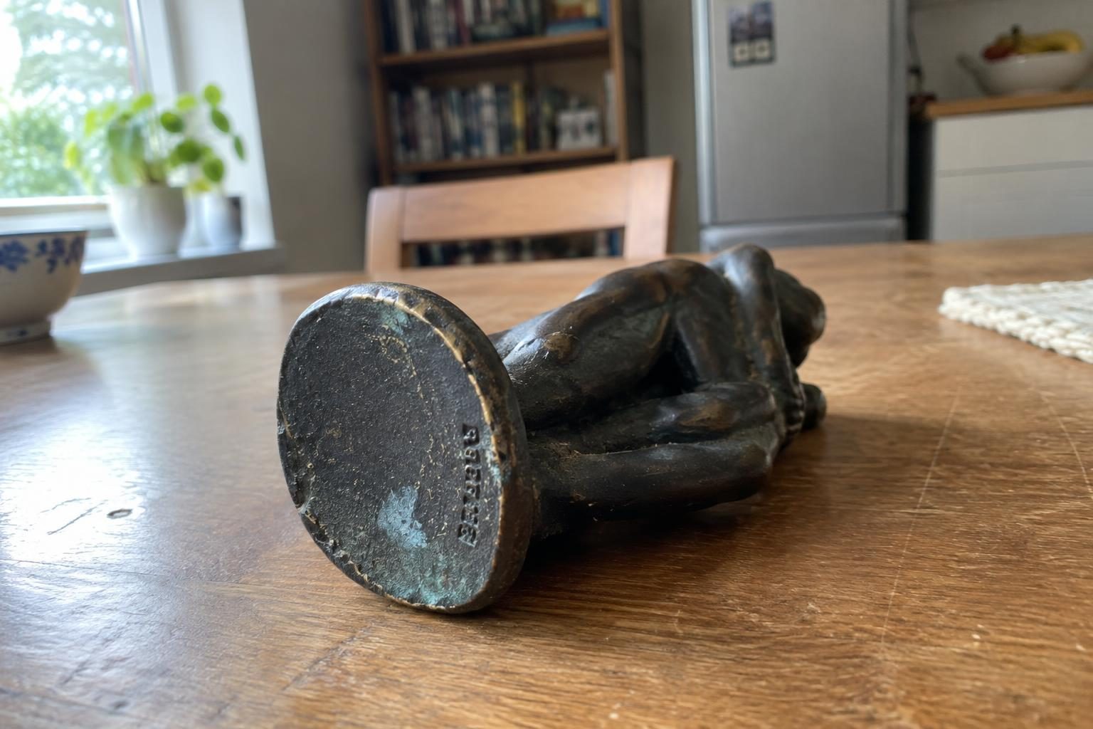 A small dark bronze figure tipped on its side on a table with faint cast lettering along the base edge