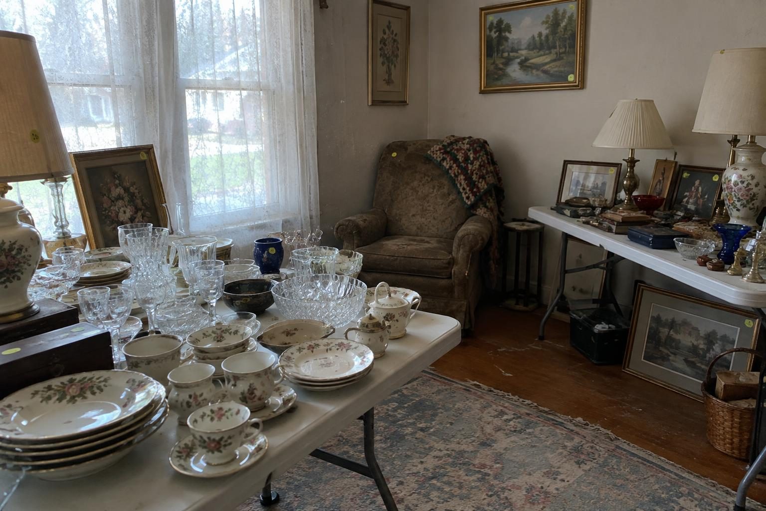 A living room set up as an estate sale, with folding tables of china, glassware, lamps and framed pictures marked with handwritten price stickers