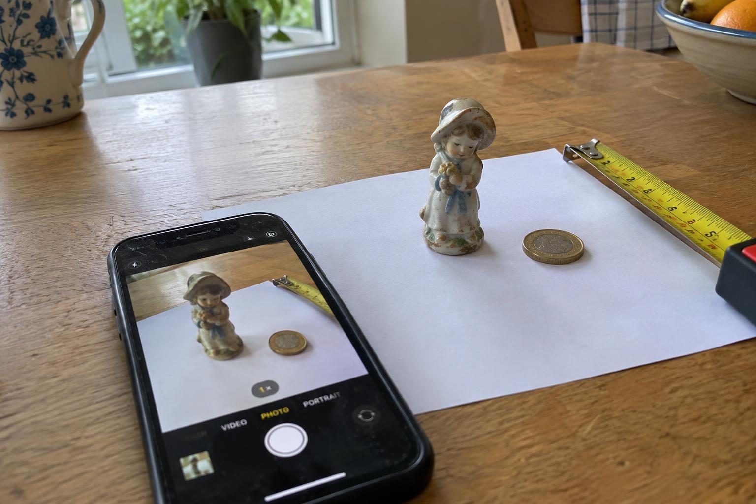 A phone photographing a small china figure on plain paper, with a tape measure and a coin beside it for scale