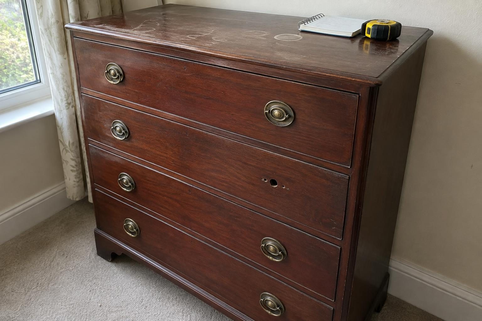 A mahogany chest of drawers in a domestic room with ring marks on the top, a missing brass handle, and a tape measure lying on it